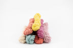 A stack of colorful yarn skeins in pastel shades including yellow, pink, purple, blue, orange, and beige, photographed on a clean white background.