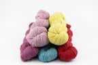 Four skeins of naturally dyed yarn in pastel and bright colors, including lavender, pale yellow, teal blue, and red, arranged in a small pile on a white background.