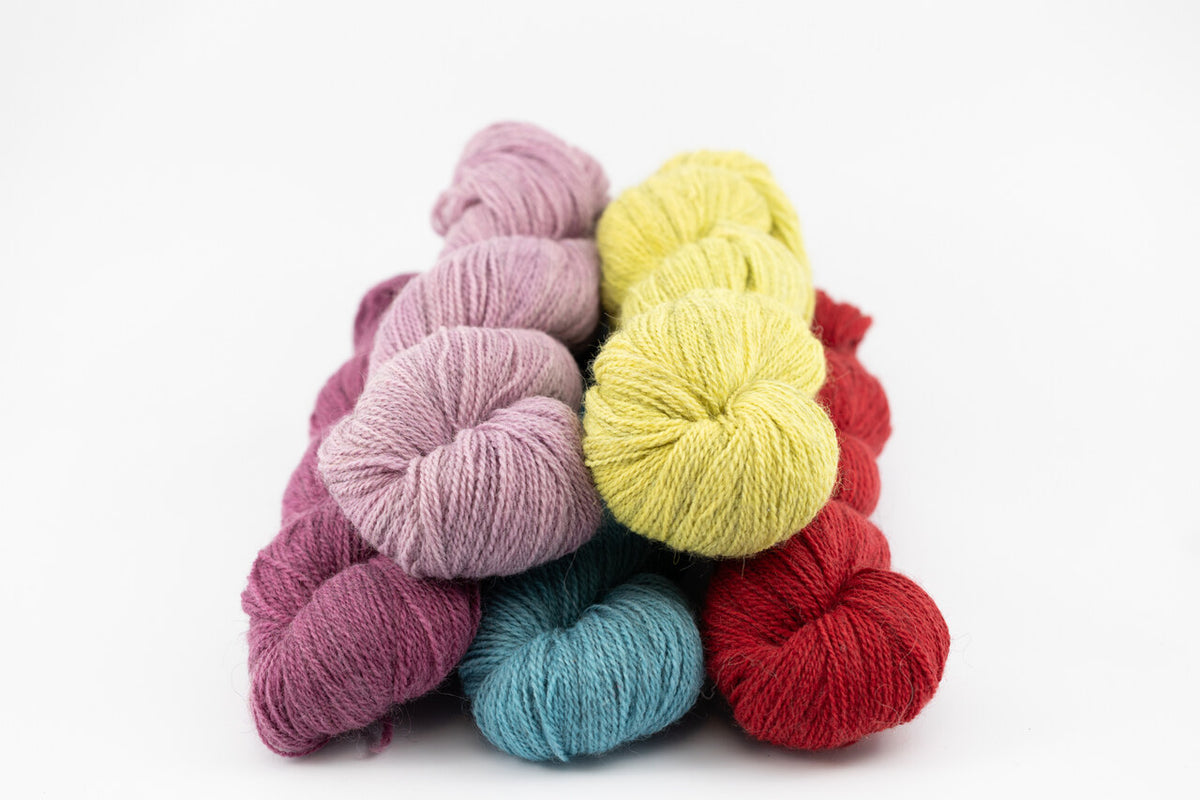 Four skeins of naturally dyed yarn in pastel and bright colors, including lavender, pale yellow, teal blue, and red, arranged in a small pile on a white background.
