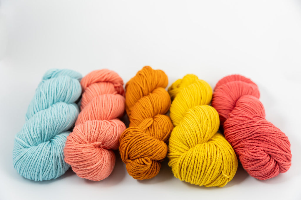 Five skeins of yarn in pastel and bright colors arranged in a row on a white background. The colors from left to right are light blue, peach, orange-brown, yellow, coral pink, and a deeper red-pink.