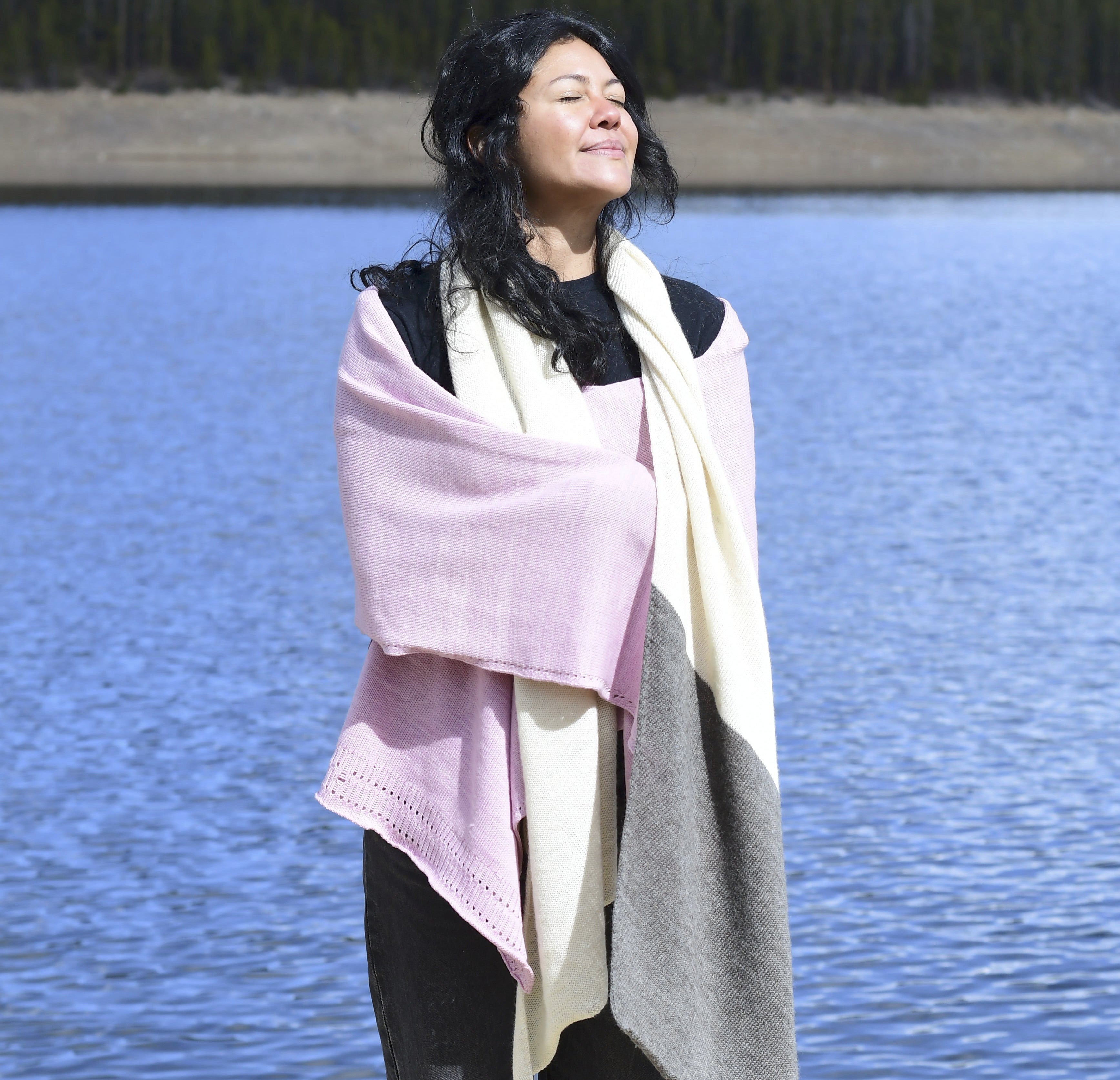 A woman standing outdoors by a lake, wrapped in wildfleece knitwear, with the blue water and forest in the background.