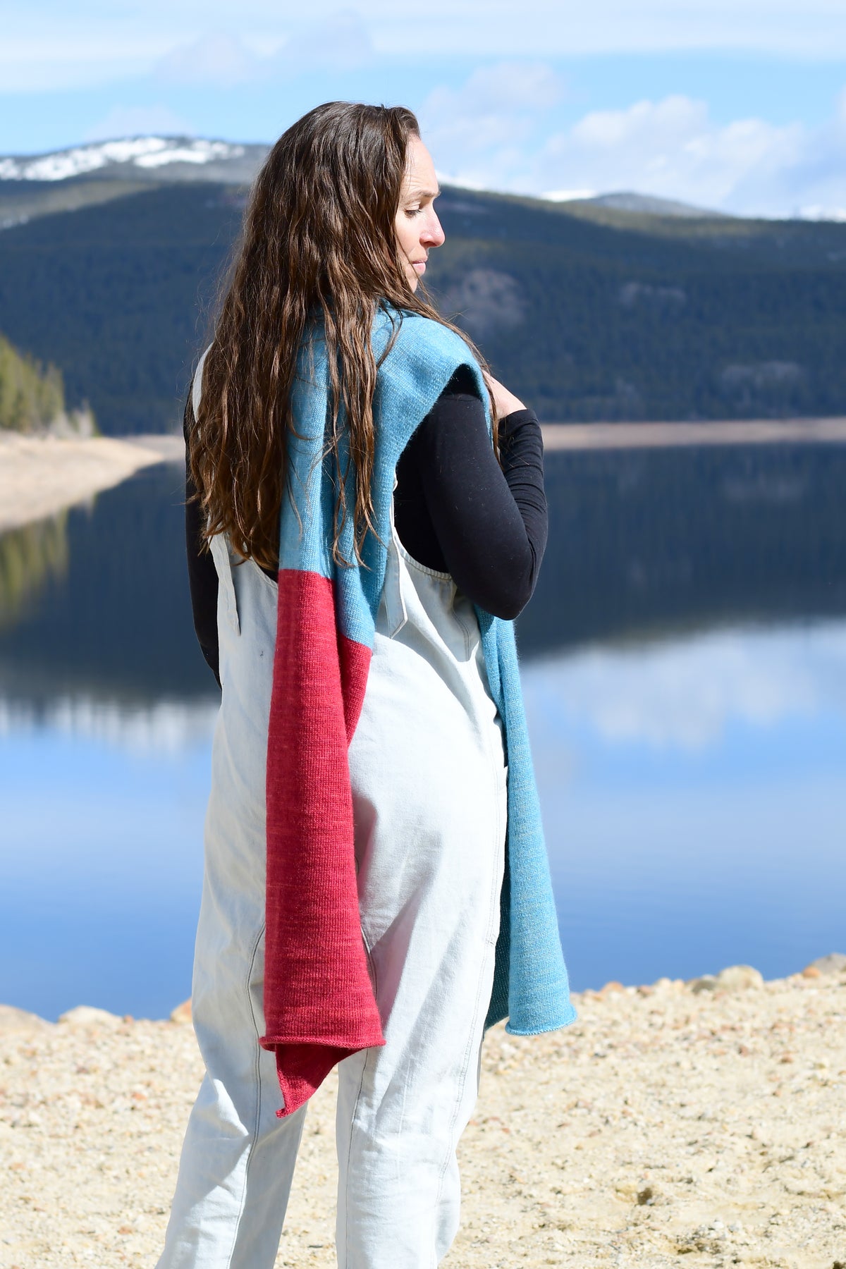 A woman standing outdoors by a lake with mountains in the background, holding a knit wrap or shawl in light blue and red.