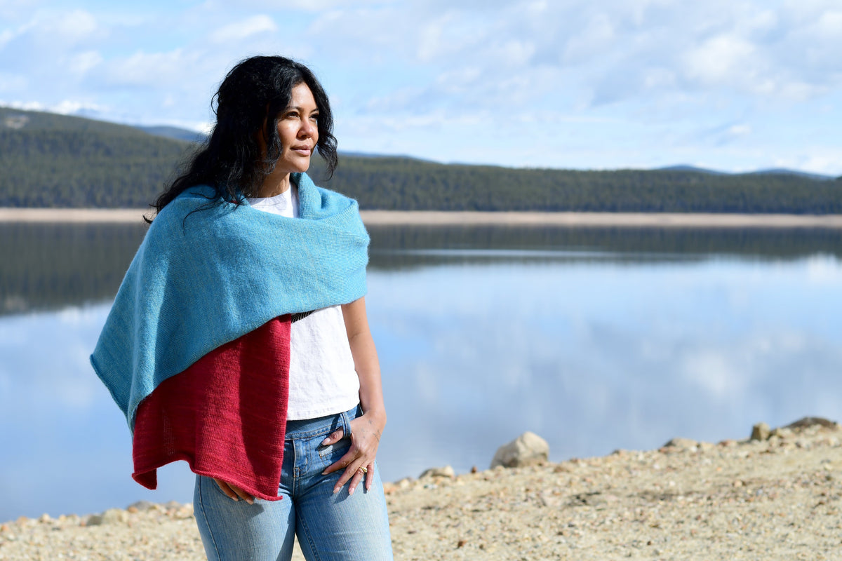 A woman standing outdoors by a lake with mountains in the background, holding a knit wrap or shawl in light blue and red.