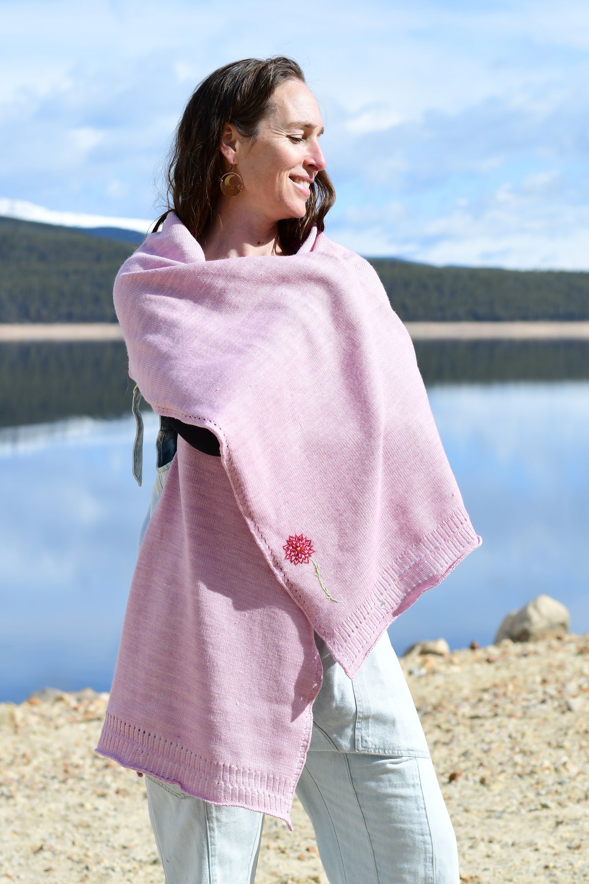 A woman wearing a light pink knitted wrap with a small red flower detail, standing outdoors by a lake with mountains in the background.
