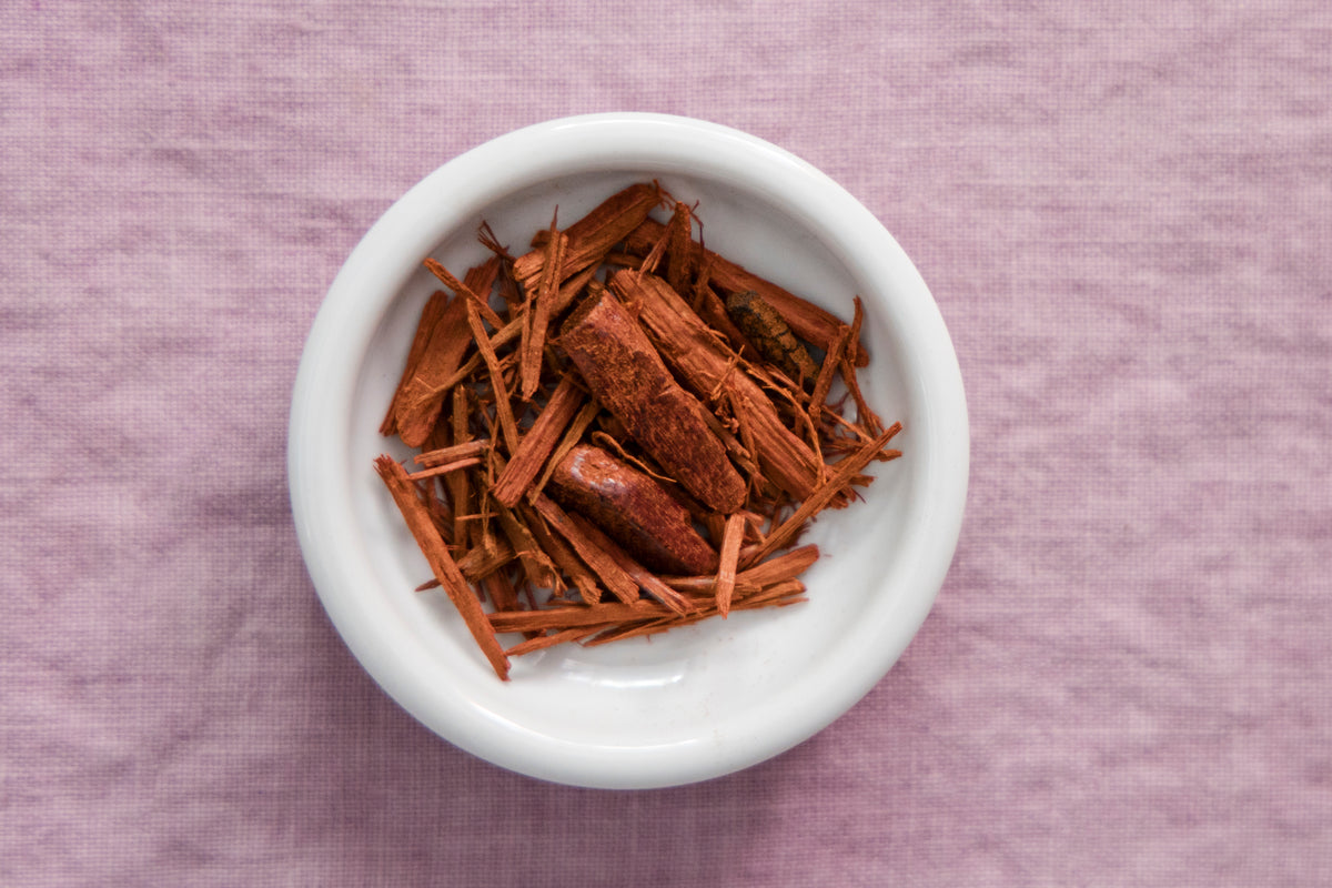 A white ceramic bowl filled with dried madder root set on a textured light purple fabric background.