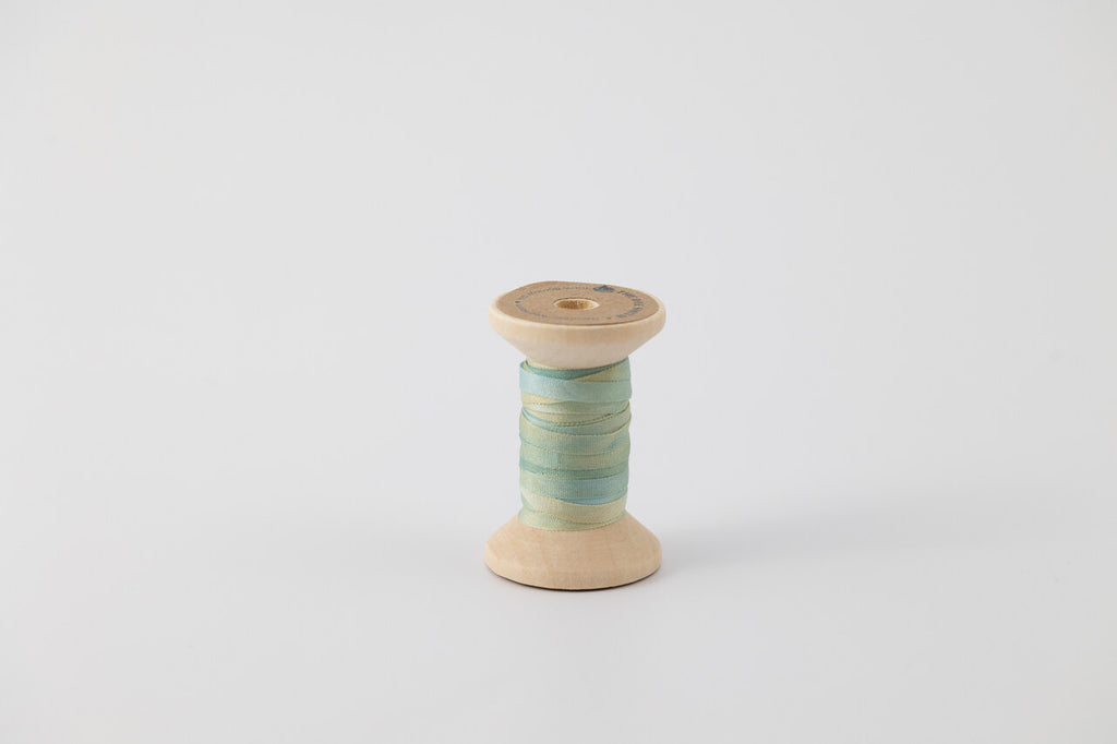 A spool of naturally dyed silk ribbon dyed with indigo and pomegranate. Multi colored with blue, green, and yellow. 