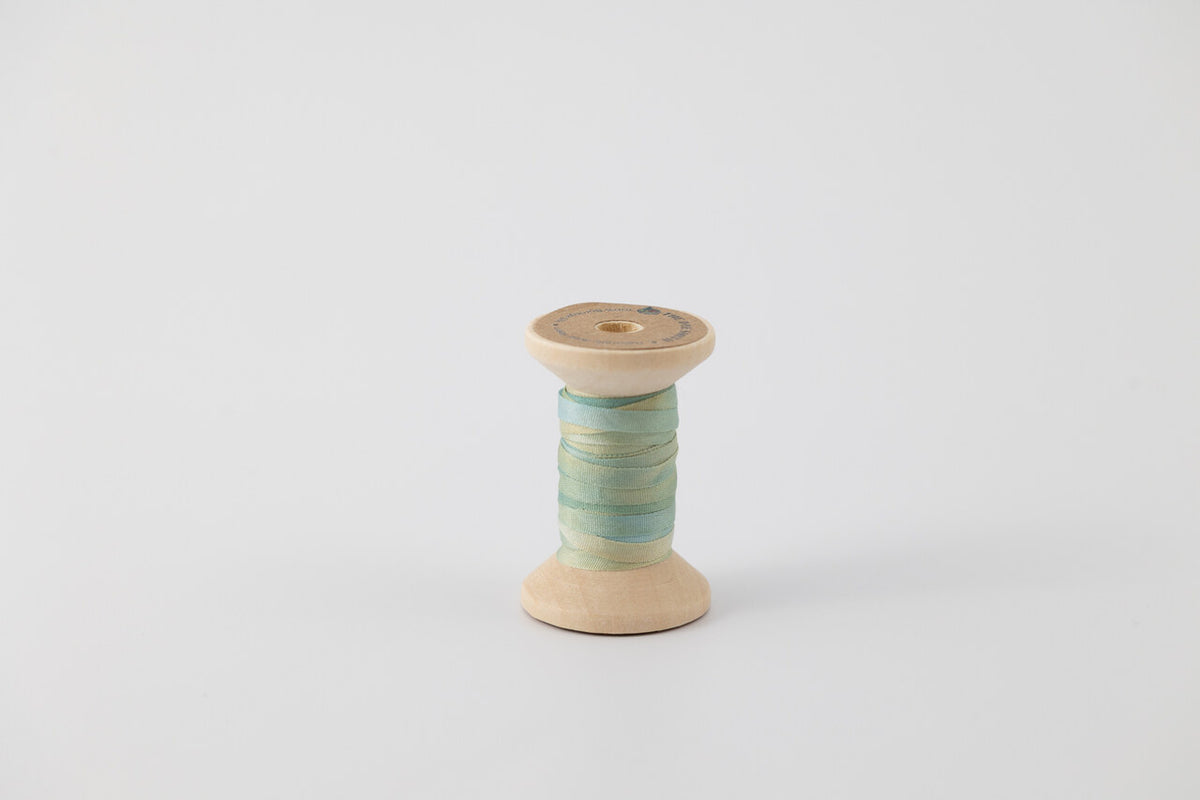 A spool of naturally dyed silk ribbon dyed with indigo and pomegranate. Multi colored with blue, green, and yellow. 