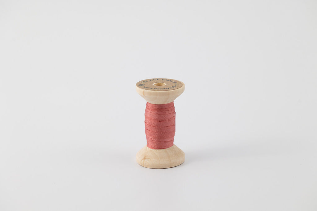 A spool of naturally dyed silk ribbon dyed with madder to a dark coral color.