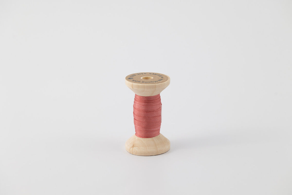 A spool of naturally dyed silk ribbon dyed with madder to a dark coral color.