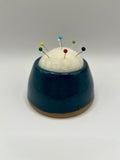 A ceramic pin holder with a dark teal blue glazed body, a natural brown unglazed base, and a white felt top holding several colorful sewing pins, set against a neutral gray background.