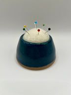A ceramic pin holder with a dark teal blue glazed body, a natural brown unglazed base, and a white felt top holding several colorful sewing pins, set against a neutral gray background.