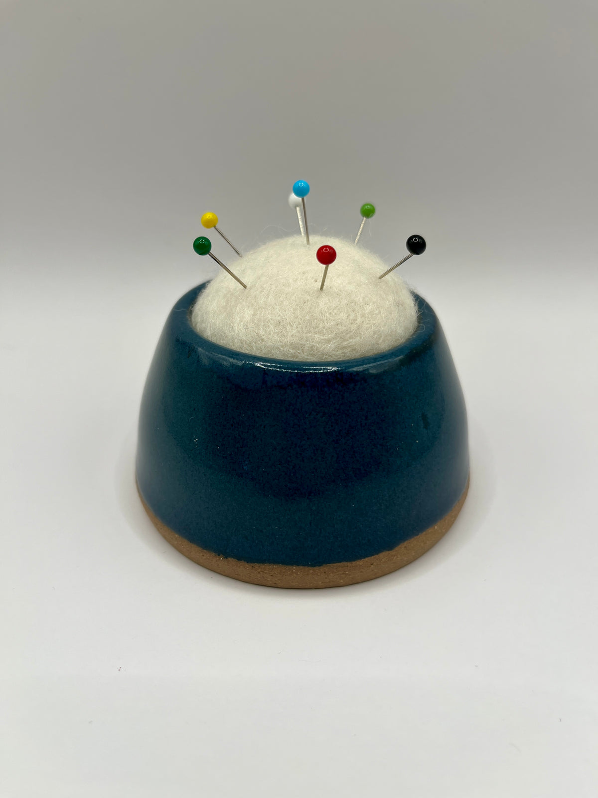 A ceramic pin holder with a dark teal blue glazed body, a natural brown unglazed base, and a white felt top holding several colorful sewing pins, set against a neutral gray background.