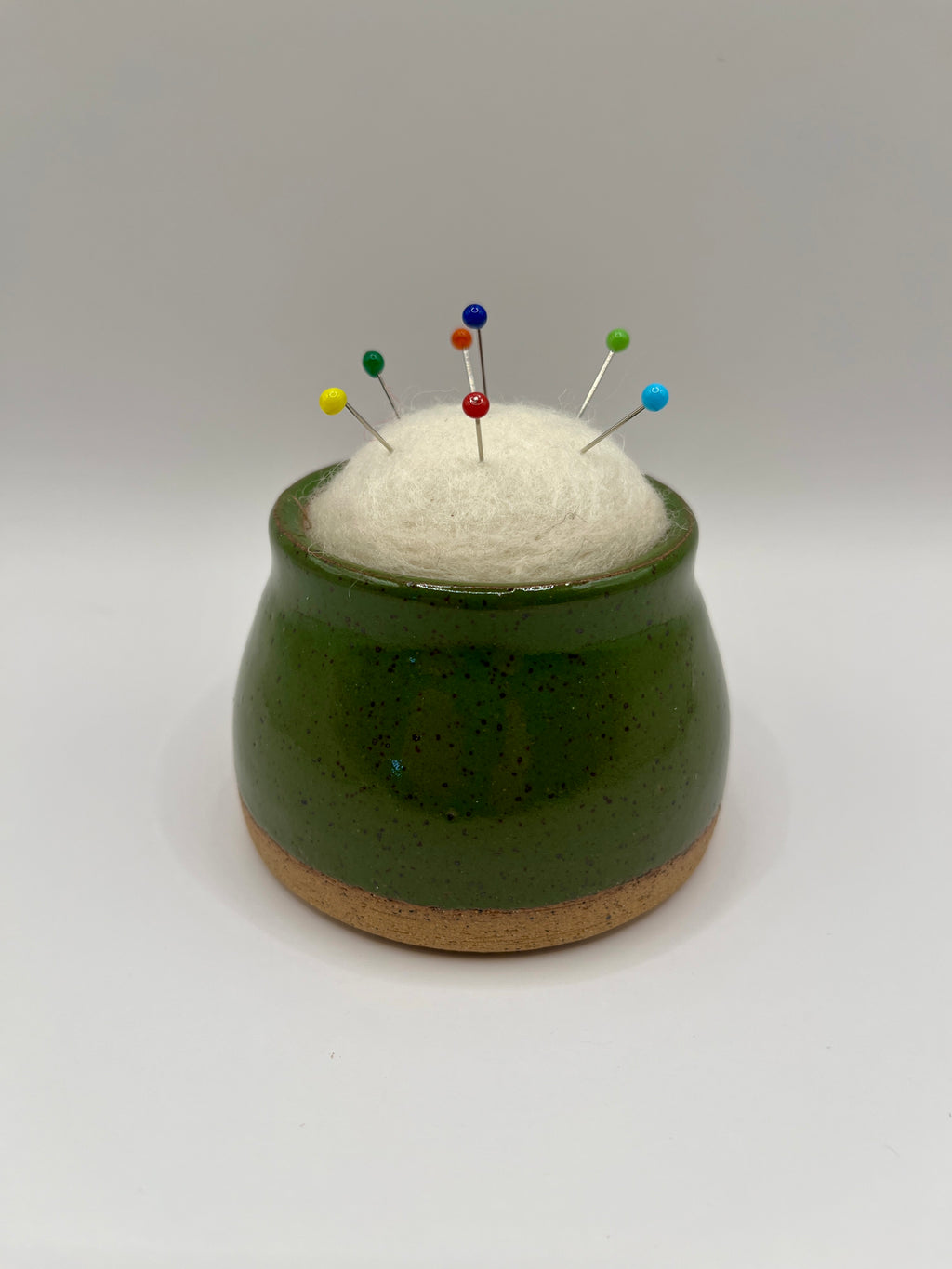 A small green ceramic pincushion with a cork base, filled with white felt and holding several colorful sewing pins, set against a plain light gray background.