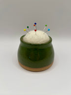 A small green ceramic pincushion with a cork base, filled with white felt and holding several colorful sewing pins, set against a plain light gray background.