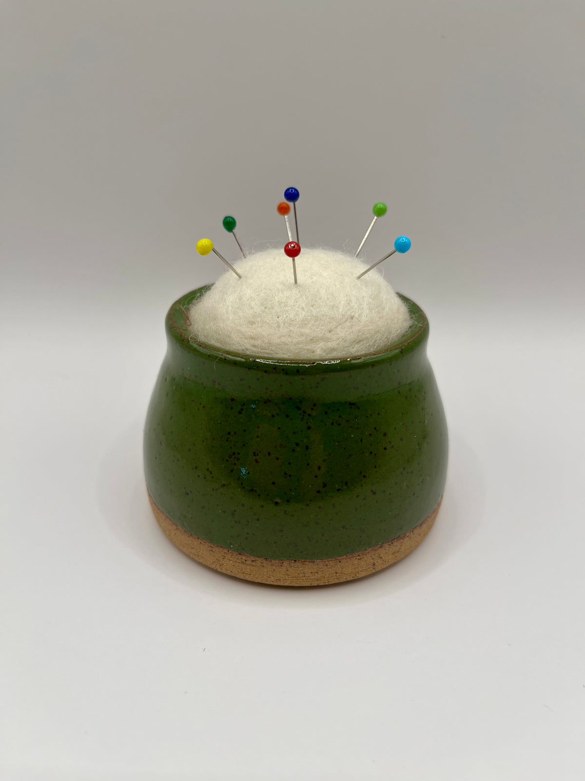 A small green ceramic pincushion with a cork base, filled with white felt and holding several colorful sewing pins, set against a plain light gray background.