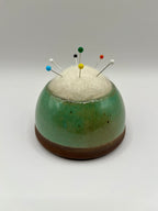 A ceramic pin holder with a rounded dome shape, featuring a green and brown glazed body, a white felt top, and several colorful sewing pins inserted into it, set against a neutral gray background.