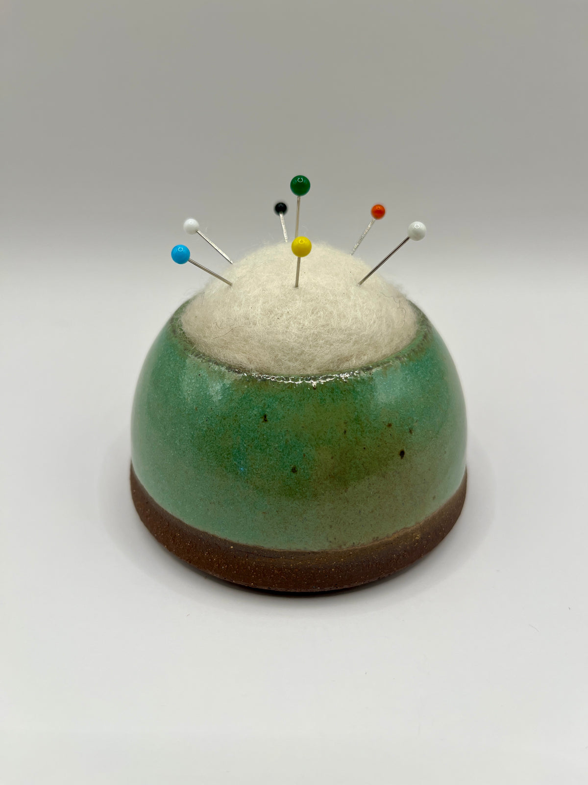 A ceramic pin holder with a rounded dome shape, featuring a green and brown glazed body, a white felt top, and several colorful sewing pins inserted into it, set against a neutral gray background.