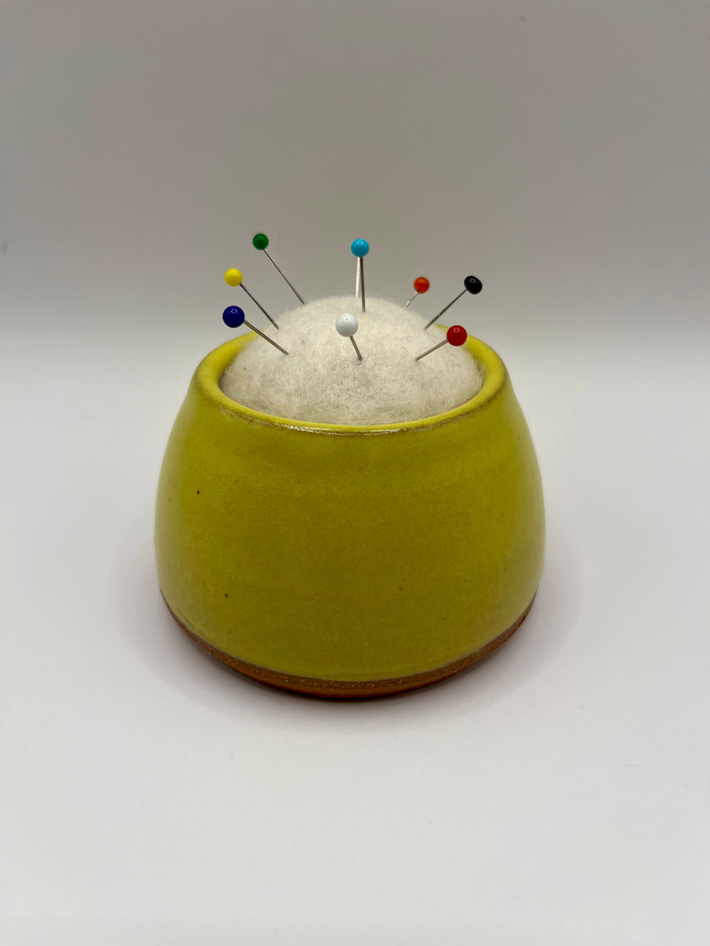 A small round yellow ceramic pin holder with a white felt top, holding several colorful sewing pins, set against a plain light gray background.