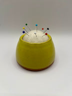 A small round yellow ceramic pin holder with a white felt top, holding several colorful sewing pins, set against a plain light gray background.