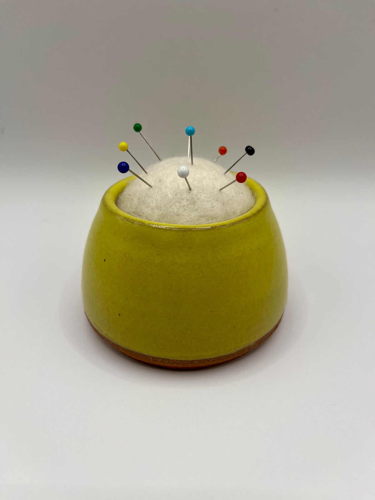 A small round yellow ceramic pin holder with a white felt top, holding several colorful sewing pins, set against a plain light gray background.