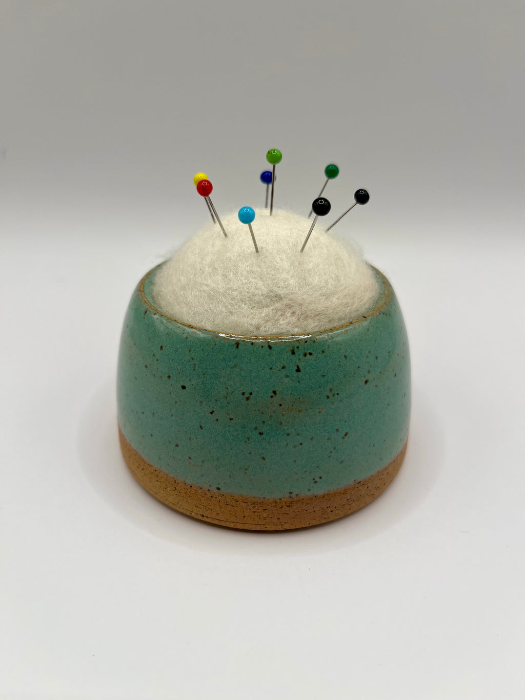 A small round ceramic pin holder with a speckled teal upper half and a natural tan clay base, filled with white cotton batting and topped with colorful sewing pins, photographed on a light gray studio background.
