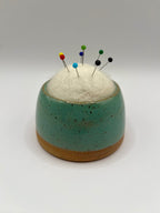 A small round ceramic pin holder with a speckled teal upper half and a natural tan clay base, filled with white cotton batting and topped with colorful sewing pins, photographed on a light gray studio background.
