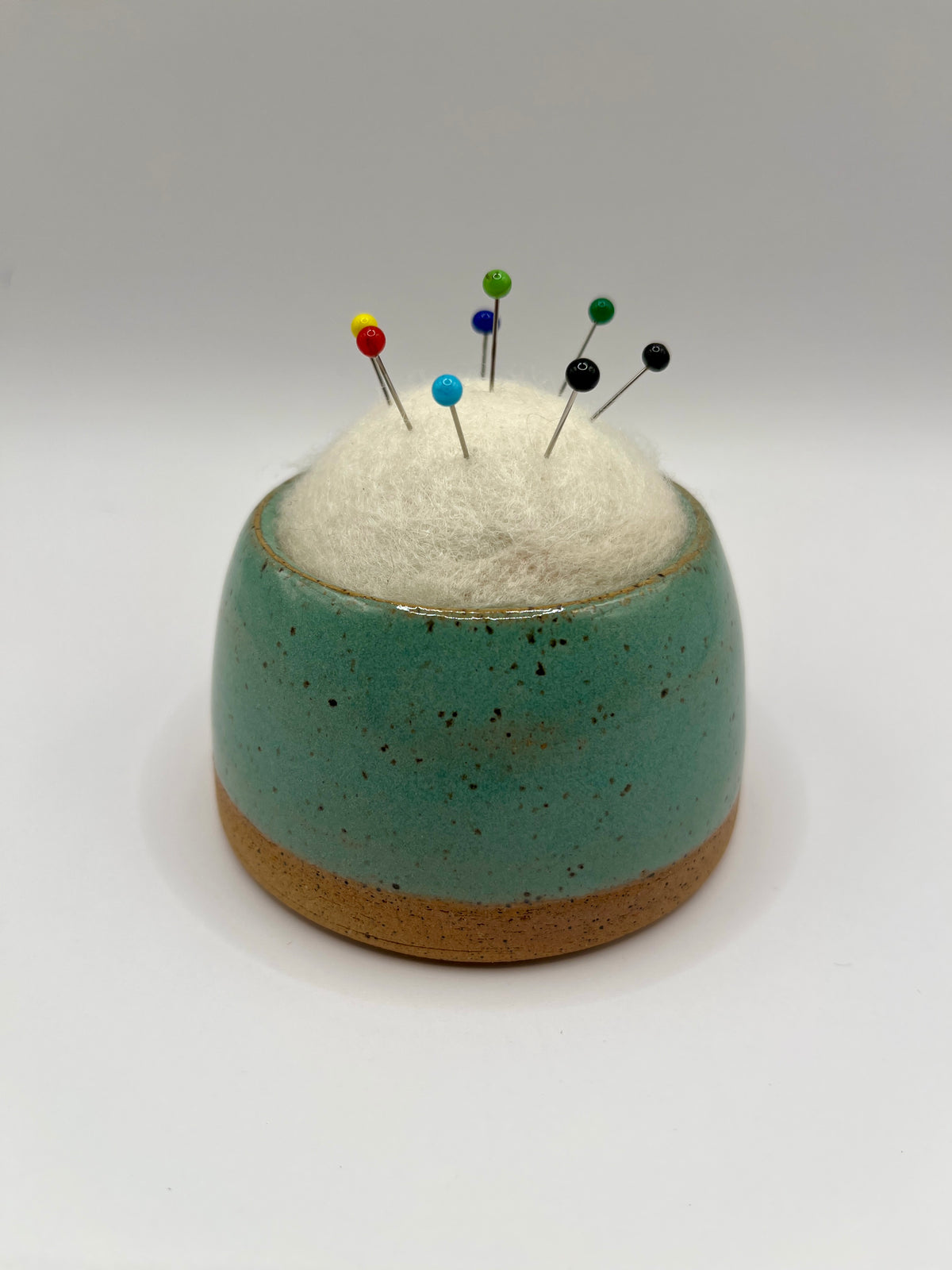 A small round ceramic pin holder with a speckled teal upper half and a natural tan clay base, filled with white cotton batting and topped with colorful sewing pins, photographed on a light gray studio background.