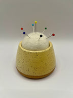 A ceramic pincushion with a speckled yellow body and a white felt pincushion on top, holding several colorful sewing pins, set against a neutral gray background.