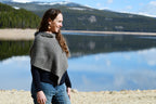 A woman wearing a gray knit poncho over a black long-sleeve top and blue jeans stands on a rocky lakeshore with mountains and water in the background.