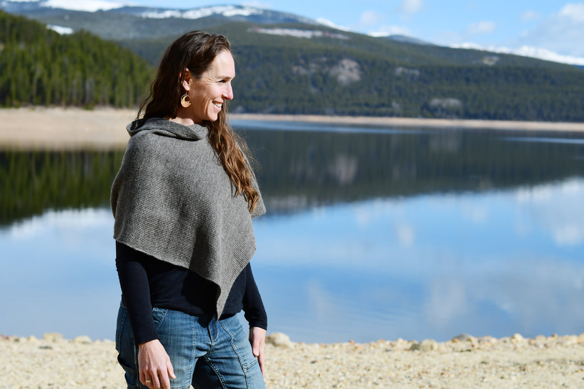 A woman wearing a gray knit poncho over a black long-sleeve top and blue jeans stands on a rocky lakeshore with mountains and water in the background.