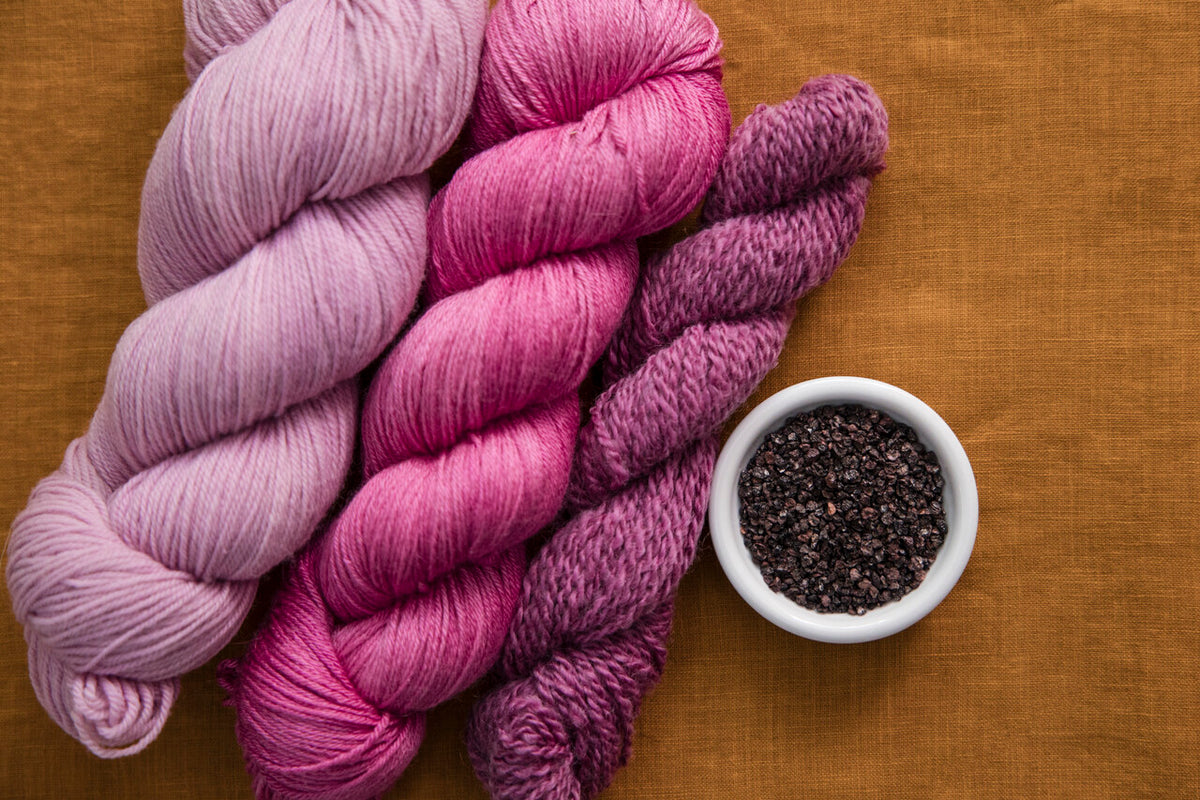 Three skeins of yarn in soft pink, magenta, and plum purple tones arranged on a warm mustard-brown fabric background, with a small white bowl of cochineal at the side.