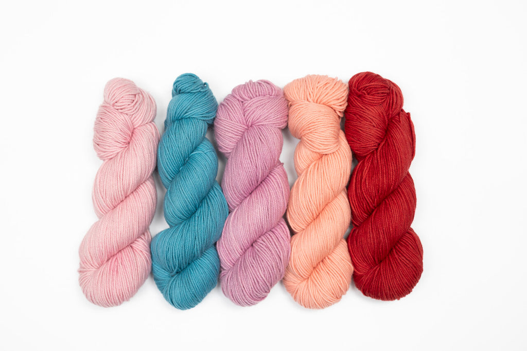 Five twisted skeins of yarn in pastel and bright shades, including pink, teal blue, lavender, peach, and deep red, arranged side by side on a plain white background.