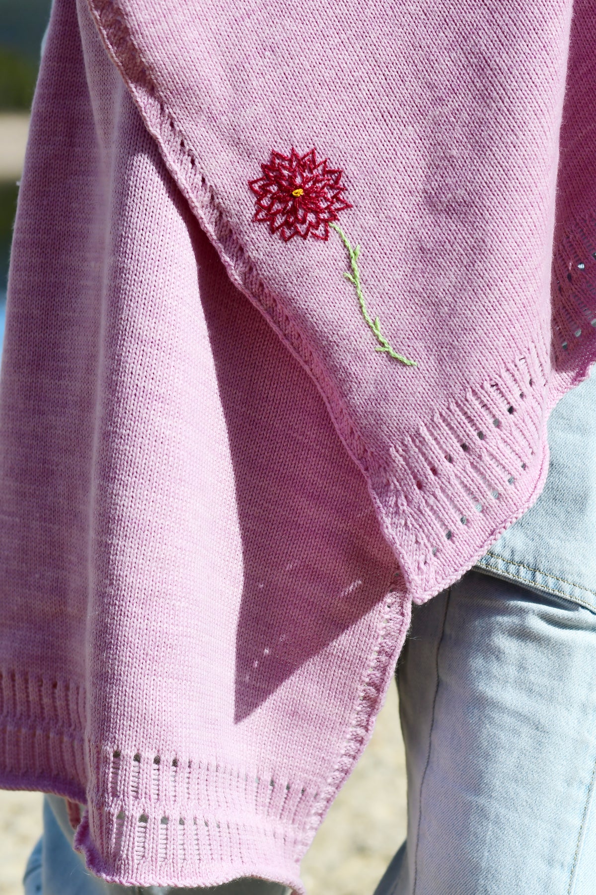 A close-up of a pink knit poncho or shawl draped over a person's shoulder, featuring a small red and green embroidered flower detail, paired with light blue jeans.